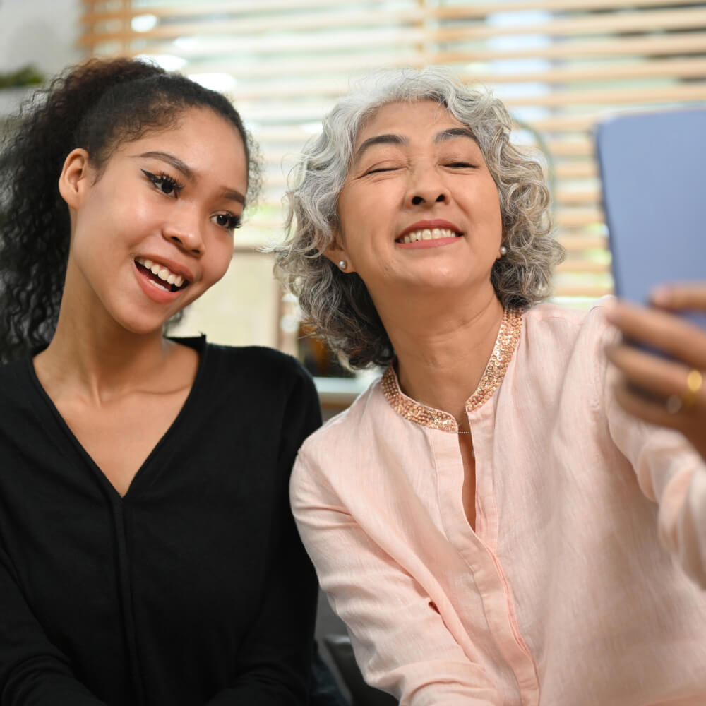 A senior woman and her granddaughter smile together at a phone screen at home.