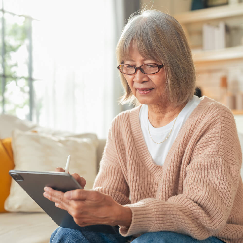 A senior woman in a cozy cardigan sketches on her tablet at home, connected and at ease.